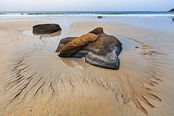 Natural rock formations resting on wet sand with gentle waves and calm sea horizon in soft daylight.