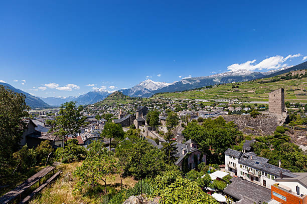 Panoramic view of Sion old town with historic buildings, vineyards, and surrounding Alpine mountains under clear blue sky.