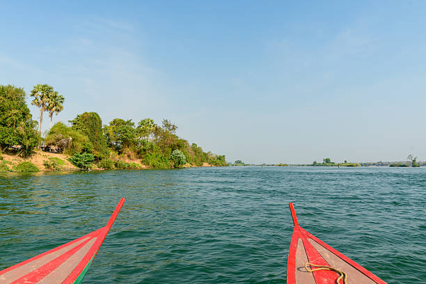 Two pointed red wooden boat prows frame a wide stretch of the Mekong River in the 4000 Islands region of southern Laos. Clear daylight reveals rippling blue water, tree-lined riverbanks, and scattered green islets beneath an open sky.