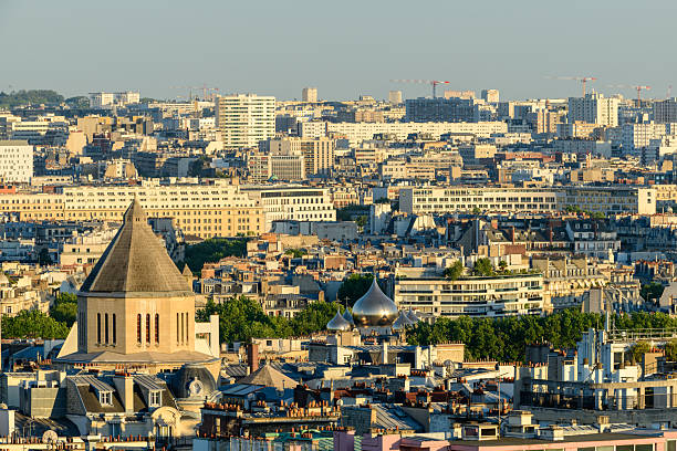 Elevated cityscape featuring the tower of Saint-Pierre-de-Chaillot and the shimmering domes of the Russian Orthodox Cathedral, with layers of Parisian buildings bathed in warm evening light.