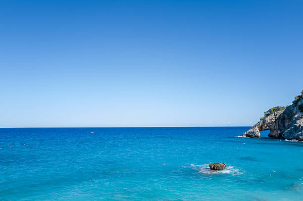 Crystal-clear turquoise sea stretches beneath a vast blue sky, with a rugged rocky headland and a lone rock emerging from the water near Cala Goloritze, Baunei, Sardinia. The scene is sunlit and tranquil, evoking a sense of openness and calm.