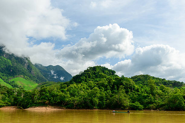 Expansive view of the Nam Ou River winding past lush green hills and dense tropical forest in northern Laos. Dramatic clouds drift above the vibrant landscape, with a small boat on the muddy water adding scale and a sense of remote tranquility.