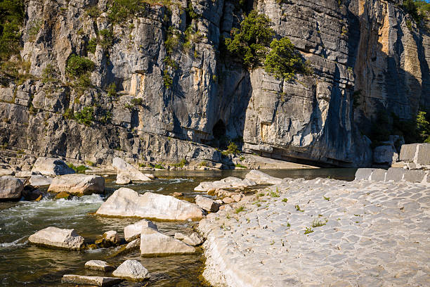 Bright sunlight illuminates a textured stone riverbank and scattered white rocks beside a dramatic vertical limestone cliff in the Gorges de l'Ardeche.