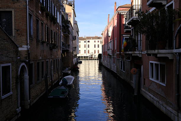View along a narrow canal lined with historic residential buildings in Venice, Italy. Sunlight reflects on the water surface while small boats are moored along the canal, illustrating everyday urban life and traditional Venetian architecture.