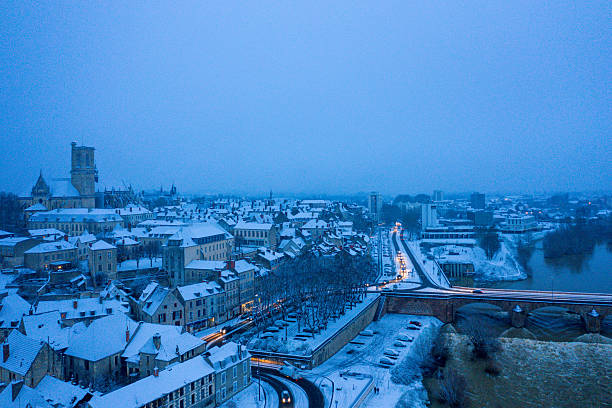 Elevated view of Nevers, France, at twilight, featuring snow-covered rooftops, historic architecture, and illuminated streets beside the Loire river in a serene winter atmosphere.
