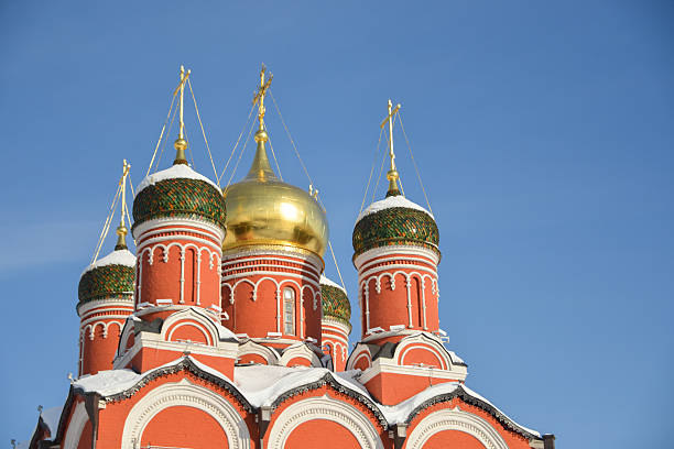 Znamensky Cathedral stands with its red-brick facade and golden domes, as viewed from Zaryadye Park in Moscow. The well-maintained landscaping around the church includes trimmed bushes and paved pathways. The cathedral’s ornate architecture is complemented by a backdrop of modern buildings, showing the contrast between historical and contemporary Moscow. Blue sky with clouds is on the background.
