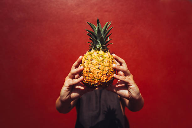 Close-up of two hands holding a ripe pineapple against a red textured background, with the fruit centered and the person partially visible.