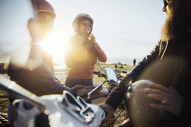 Group of motorcyclists wearing helmets and leather jackets talking beside their motorcycles outdoors, backlit by warm sunset light with a coastal landscape in the background.