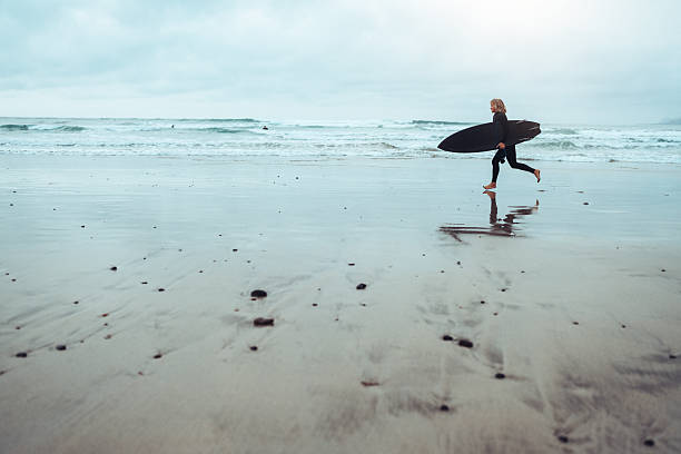 Female surfer in a wetsuit running barefoot along a wet, reflective beach while carrying a surfboard toward the ocean. Moody coastal scene with soft light and misty background conveying freedom, solitude, surf lifestyle, and connection with nature.
