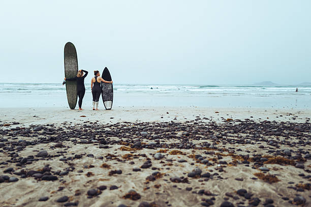 istock Female Surfers Standing on Beach Facing the Ocean 2255726373
