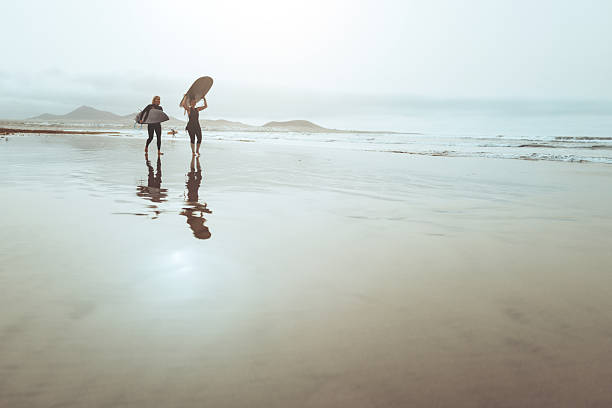 Two female surfers in wetsuits carry surfboards while walking along a sandy beach near the ocean. Natural coastal scene capturing surf lifestyle, outdoor sport, friendship, and anticipation before entering the water.