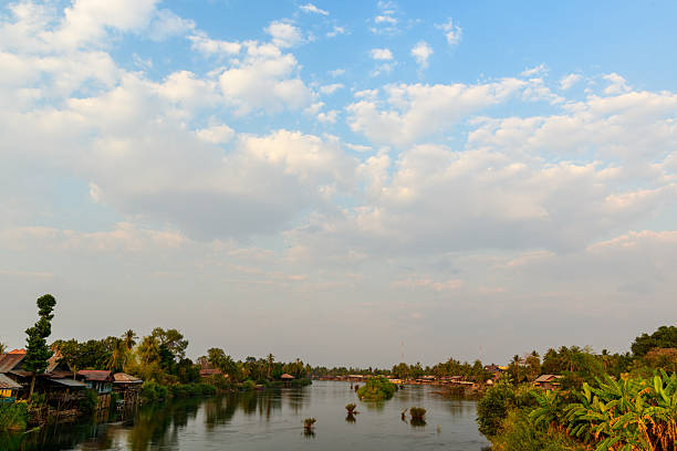 Traditional wooden houses and lush tropical vegetation line the banks of the Mekong River in the 4000 Islands region of southern Laos. Scattered islets and a wide expanse of sky with soft clouds create a tranquil riverside scene in gentle daylight.