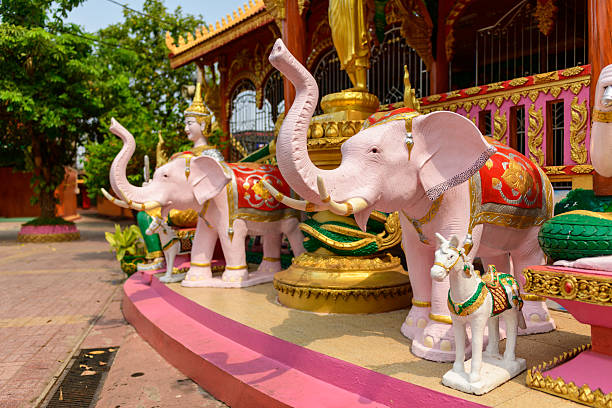 Ornate pink elephant sculptures with gold and red detailing stand beside a small white horse figure in the sunlit courtyard of Vat Simuong temple, Vientiane. Traditional architectural elements and lush greenery frame the vibrant scene.