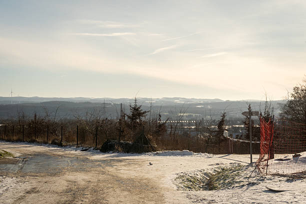 Snowy dirt road stretching through a peaceful rural landscape. A fence lines the path, with distant hills and soft winter sunlight, capturing the serenity of a winter countryside escape.