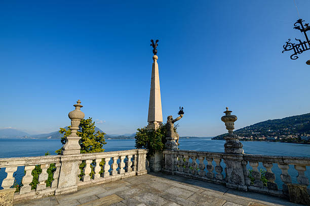 A tall stone obelisk and classical statue rise above an ornate balustrade terrace overlooking the blue waters of Lake Maggiore in the historic Borromean Gardens, Italy. Crisp sunlight and clear sky highlight elegant garden architecture and panoramic mountain scenery.