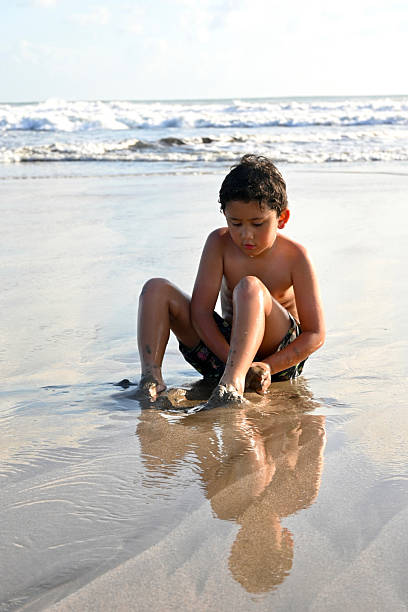 Boy playing in shallow water at beach, summer holidays. Child squatting near waves, enjoying sensory play with wet sand at seaside.