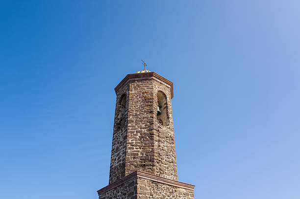Octagonal stone bell tower with arched openings stands against a clear blue sky in Castelsardo, Sardinia. Sunlight highlights the rough masonry and architectural details.