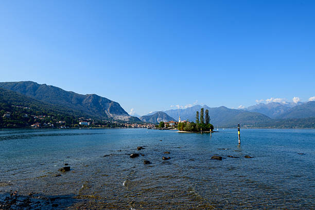 Wide view of Isola Bella surrounded by the blue waters of Lake Maggiore, with distant alpine mountains and lakeside villages under a bright sky. The tranquil scene features lush greenery and gentle ripples along the shoreline in northern Italy.