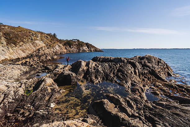 Person wearing outdoor clothing and backpack walking across rocky coastal terrain near calm water. The reflection in the shallow pool mirrors the figure against a clear sky, conveying exploration, balance, and connection with nature. Scenic outdoor lifestyle image with copy space.