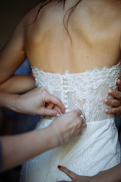 Bride is standing in front of mirror and fitting dress in atelier with wedding assistant. Woman with dressmaker are making final touch on tailor made gown in bridal clothing shop.