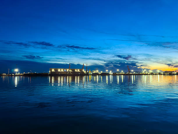 Evening at a harbor where water reflects lights from buildings and boats. Clouds are present in the sky as the sun sets, creating a colorful scene.