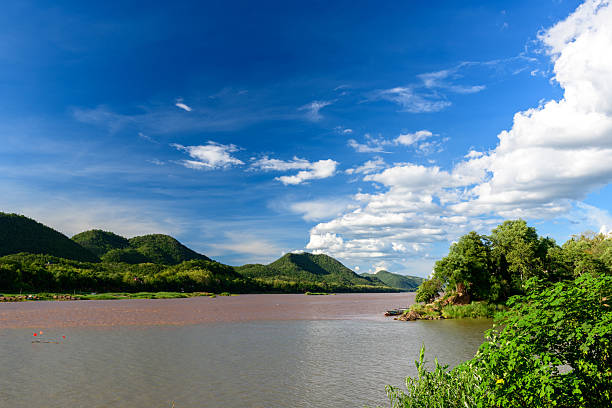 Expansive view of the muddy Mekong river winding past lush green hills and dense riverside vegetation near Luang Prabang, Laos. Bright blue sky with scattered clouds creates a vivid, natural landscape ideal for travel and nature concepts.