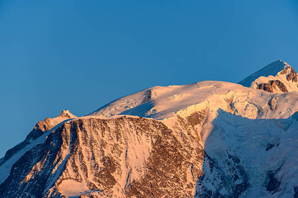 Early morning sunlight bathes the snow-covered summit and rugged rock faces of Mont Blanc in warm orange tones, set against a deep blue sky for a striking alpine contrast.