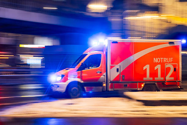 A Notruf 112 emergency ambulance from Fire Department drives through Berlin at night with blue warning lights flashing, photographed close-up with motion blur and urban light streaks.