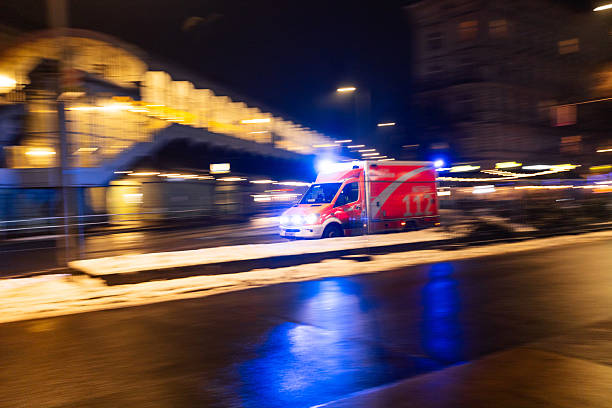A Berlin emergency ambulance marked 112 moves through the city at night with blue lights flashing, captured with panning blur, snow along the roadside, and reflections on the street.