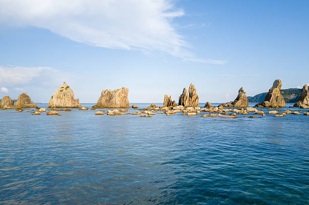 Distinctive rocky pillars emerge from calm blue coastal waters beneath a bright sky at Hashiguiiwa, Kushimoto, creating a striking natural seascape with textured stone and gentle sunlight.