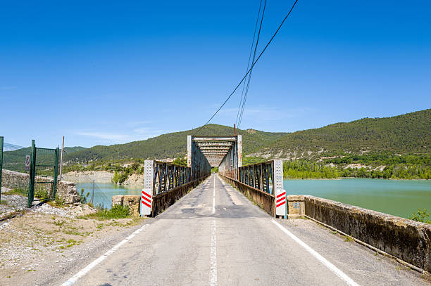 A narrow metal truss bridge stretches across calm green reservoir waters, framed by rugged hills and forest under a clear blue sky at Las Penas. The scene features strong linear perspective and vibrant spring colors.