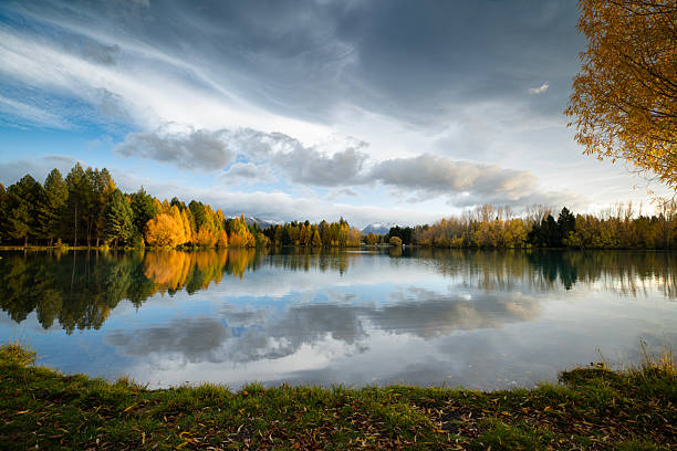 A dramatic cloudscape fills the sky above Lake Ruataniwha. The early morning sun breaks through the clouds to shine on the golden autumn landscape. The lake is still and reflects the scene. In the foreground, golden leaves have fallen onto the grassy bank. Lake Ruataniwha is situated near Twizel, in the center of New Zealand's South Island. It is popular with both local and international tourists.