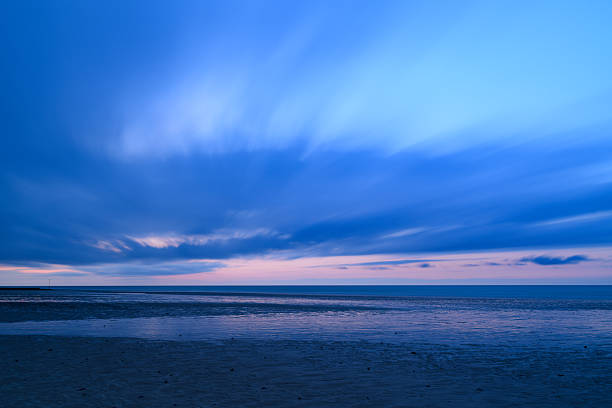 Long exposure captures streaked blue clouds and tranquil water at low tide along the English Channel, with soft pastel light fading on the horizon and wet sand reflecting the sky.