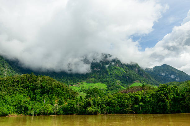 Low clouds drift across lush green hills above the muddy Nam Ou River in northern Laos. Dense tropical forest covers the slopes, creating a vibrant landscape under a bright, overcast sky.