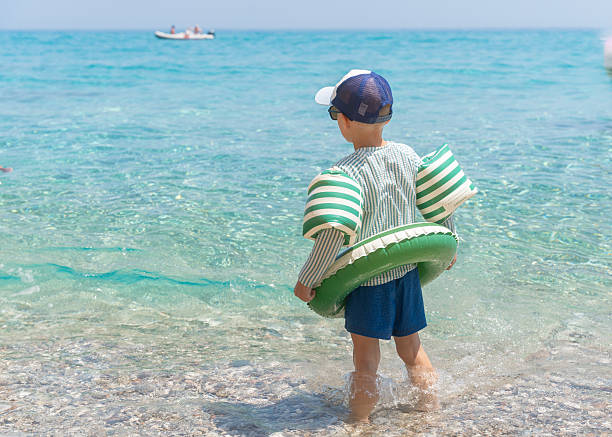 Child standing at the edge of a clear turquoise sea with rocky cliffs in the background. Bright sunlight highlights the shallow water and relaxed summer atmosphere of a quiet Mediterranean beach in Sardinia, Italy. Cala luna