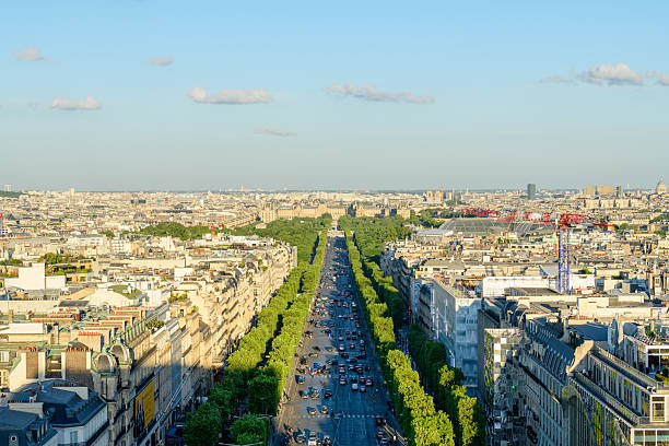 High angle view of the Avenue des Champs-Elysees lined with vibrant green trees, stretching through central Paris under a clear blue sky with scattered clouds and evening sunlight.