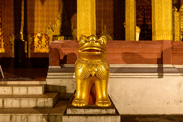 A golden guardian lion statue stands at the entrance of an ornately decorated Buddhist temple in Luang Prabang, Laos. Warm artificial lighting highlights intricate patterns and rich architectural details in this traditional Southeast Asian setting.