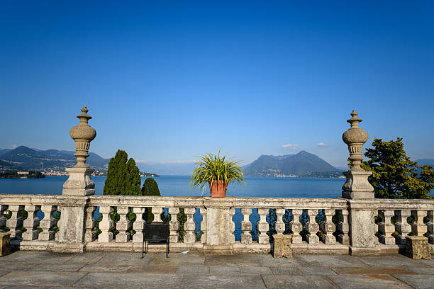 Classic stone balustrade terrace with ornamental urns and a potted plant offers an open view across Lake Maggiore toward distant mountains under a vivid blue sky. The elegant setting features geometric lines, lush greenery, and tranquil water in northern Italy.
