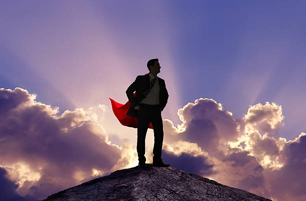 A man wearing a cape stands proudly as he is silhouetted by sunbeams emanating from behind clouds.