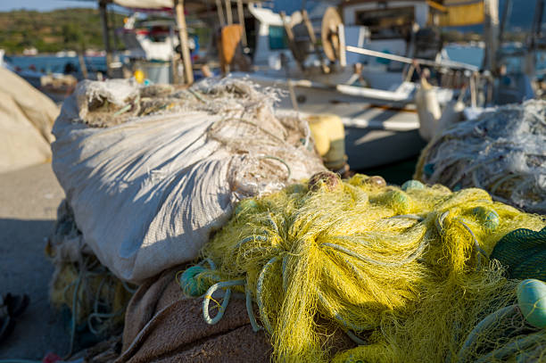 Close-up of tangled yellow fishing nets and sacks resting on a sunlit dock with blurred fishing boats in the background at a harbor in Argolide, Peloponnese.