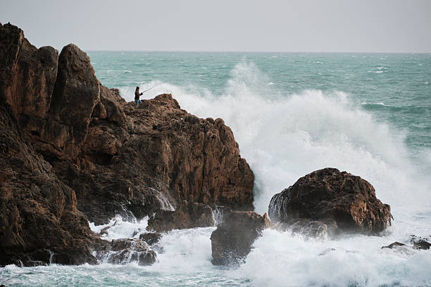 A lone person fishing from a rocky shore with massive white sea spray from waves hitting the coast.