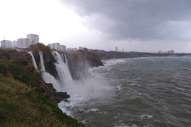 A powerful waterfall falling from a cliff into the rough sea under a gray stormy sky with city buildings in the distance.