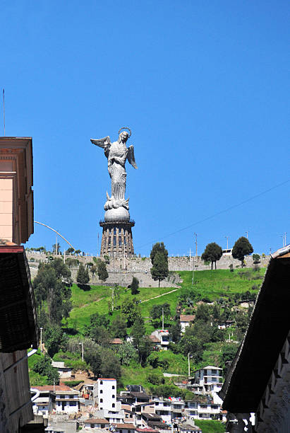 Quito, Pichincha, Ecuador: the Virgin of El Panecillo (also known as the Virgen de Quito), a monumental aluminum statue standing atop El Panecillo hill in Quito, Ecuador. Winged Virgin of the Apocalypse standing on a globe and chaining a dragon, based on the 18th-century sculpture by Bernardo de Legarda. It overlooks the historic city center, a UNESCO World Heritage Site. The image captures the statue against a clear blue sky, rising above the green slopes of the hill which are dotted with residential houses and trees. In the immediate foreground, the eaves of colonial-style roofs frame the view of the volcanic hill.