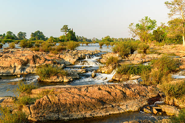 Gentle morning light highlights layered rocks, small waterfalls, and scattered greenery at Tat Somphamit Waterfalls, also known as Li Phi Falls, in Champasak Province, southern Laos. The scene features textured stone, flowing water, and lush riverbank vegetation in a tranquil natural setting.