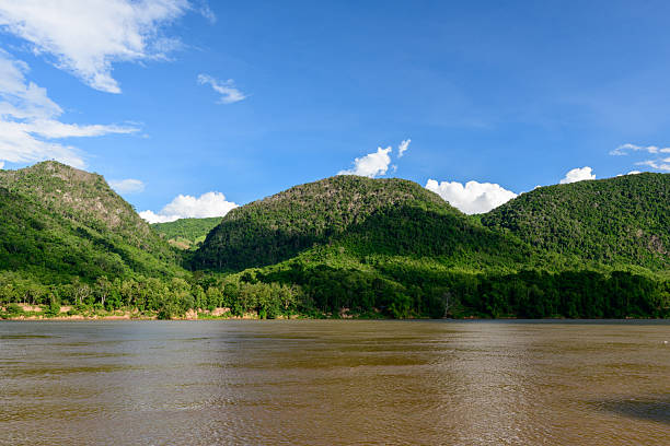 The Mekong river flows past densely forested hills with sunlight casting shadows across the lush slopes. A vivid blue sky with scattered white clouds enhances the tranquil natural landscape.