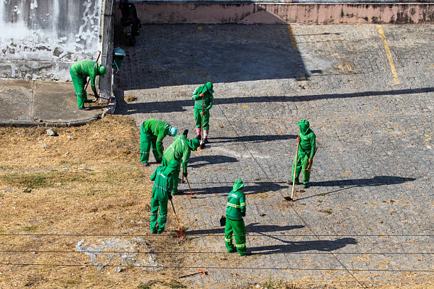 Municipal sanitation workers sweep and maintain sidewalks and public areas using brooms, shovels, and wheelbarrows. Uniformed employees work in an urban environment under natural light. The image represents municipal services, city conservation, collective work, and routine urban upkeep.