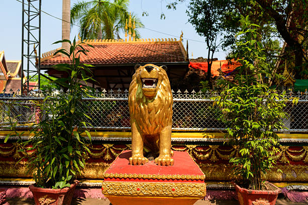 A gilded lion sculpture stands on a red pedestal in the sunlit courtyard of Vat Simuong temple, Vientiane. Ornate gold detailing, leafy potted plants, and traditional Lao architectural elements create a vibrant and decorative scene.