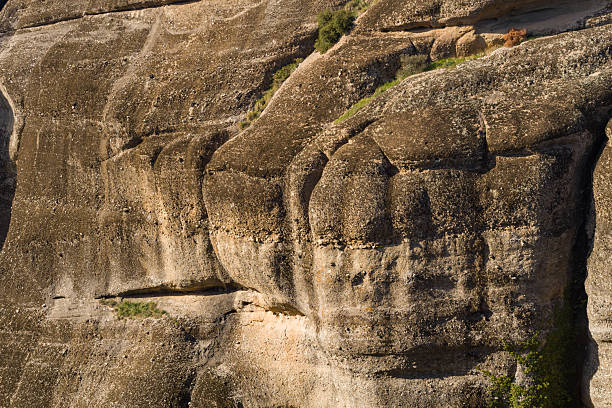 Detailed view of rugged sandstone cliff face in Meteora, Greece, showcasing layered textures, natural erosion patterns, and warm sunlight highlighting earthy tones.