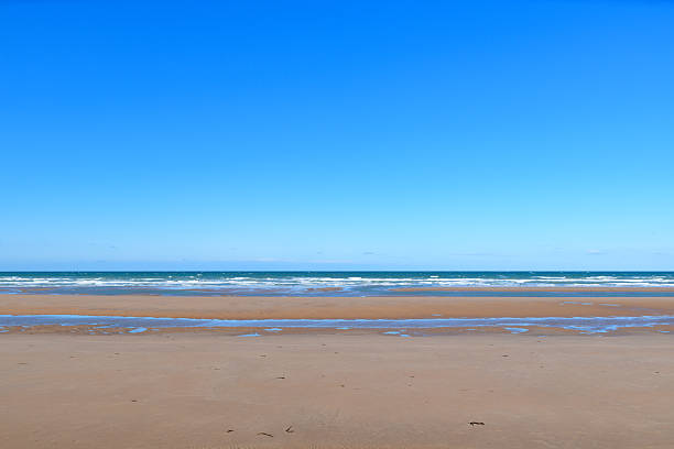 Wide sandy expanse of Omaha Beach stretches to the horizon, with gentle waves meeting the shore beneath a vibrant blue sky. The scene is open, calm, and uncluttered, evoking a sense of space and tranquility.