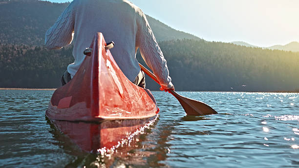 A person paddles a red canoe on a serene lake, surrounded by forested mountains. The scene captures a peaceful outdoor activity, ideal for weekend adventures and promoting an active lifestyle. The focus on the paddle and the tranquil setting highlights the connection with nature, captured with a worm's-eye view.